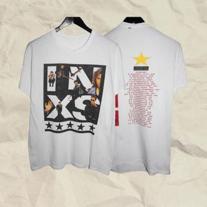 May include: A white short-sleeve t-shirt featuring a black graphic on the front with the text INXS and five stars. The back displays a yellow star above the text Calling All Nations Tour and a list of cities and dates in red and black ink.