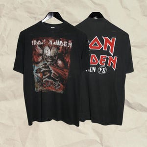 May include: A black short-sleeve t-shirt featuring an Iron Maiden graphic on the front with a skeletal figure and the text Virtual XI. The back of the shirt displays the Iron Maiden logo in red and white with the text Tour 98 in a circle.