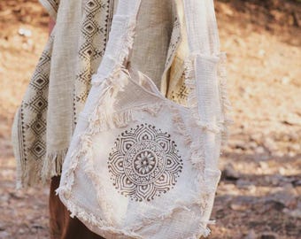 May include: Cream-colored shoulder bag with a mandala design. The bag has a fringed edge and a long strap. The mandala is printed in a dark brown color. The bag is made of a woven fabric.