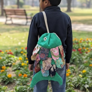 May include: A green fish-shaped backpack with colourful patterned fins is worn by a person in a park. The backpack has a drawstring closure and a white strap. The person is wearing a navy blue sweater and blue jeans.