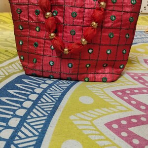 May include: Red tote bag with a square pattern, green accents, and gold beaded handles. The bag is made of a shiny fabric and sits on a colorful patterned surface. The bag's handles are made of red fabric with gold beads.