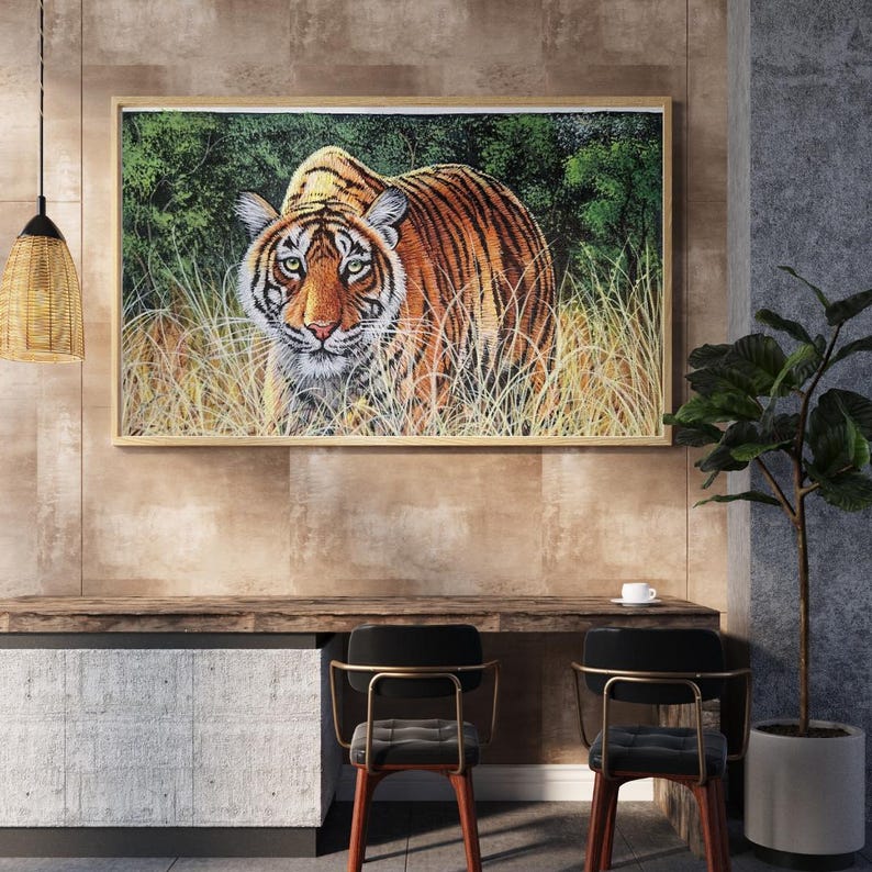 May include: A framed artwork featuring a detailed painting of a tiger in its natural habitat. The tiger has orange and black stripes, green eyes, and is surrounded by tall grass and green foliage. The artwork is displayed on a wall above a wooden desk.