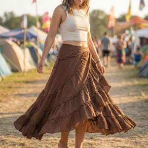 May include: A woman wearing a white crop top and a long, brown, tiered skirt. The skirt has a ruffled design. The photo was taken outdoors, with tents and people in the background. The skirt is a bohemian style.