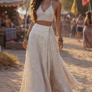 May include: A two-piece outfit consisting of a cream-coloured crochet halter crop top and a matching long, flowing skirt. The skirt features a wide, smooth waistband with side lace-up detailing and a textured, frayed fabric finish throughout.