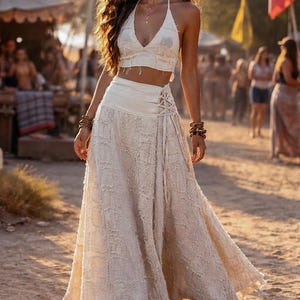 May include: A two-piece set consisting of a cream-colored halter crop top with a V-neckline and a matching high-waisted maxi skirt. The skirt features a wide waistband with side lace-up detailing and a textured, frayed fabric construction.
