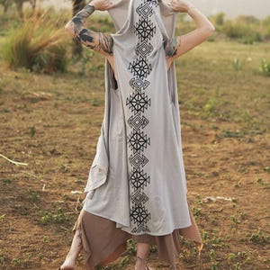 May include: A light gray sleeveless hooded vest featuring a long, vertical black geometric pattern running down the center of the back and hood. The garment is worn over a brown skirt with an uneven hemline, creating a layered appearance.