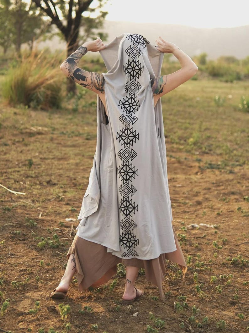 May include: A light gray sleeveless hooded vest featuring a long, vertical black geometric pattern running down the center of the back and hood. The garment is worn over a brown skirt with an uneven hemline, creating a layered appearance.