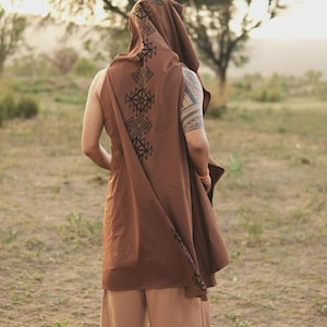 May include: A brown sleeveless hooded tunic featuring a black geometric pattern printed along the hood and down the back. The garment is worn with light brown trousers. The tunic has a loose, flowing fit and a pointed hood design.