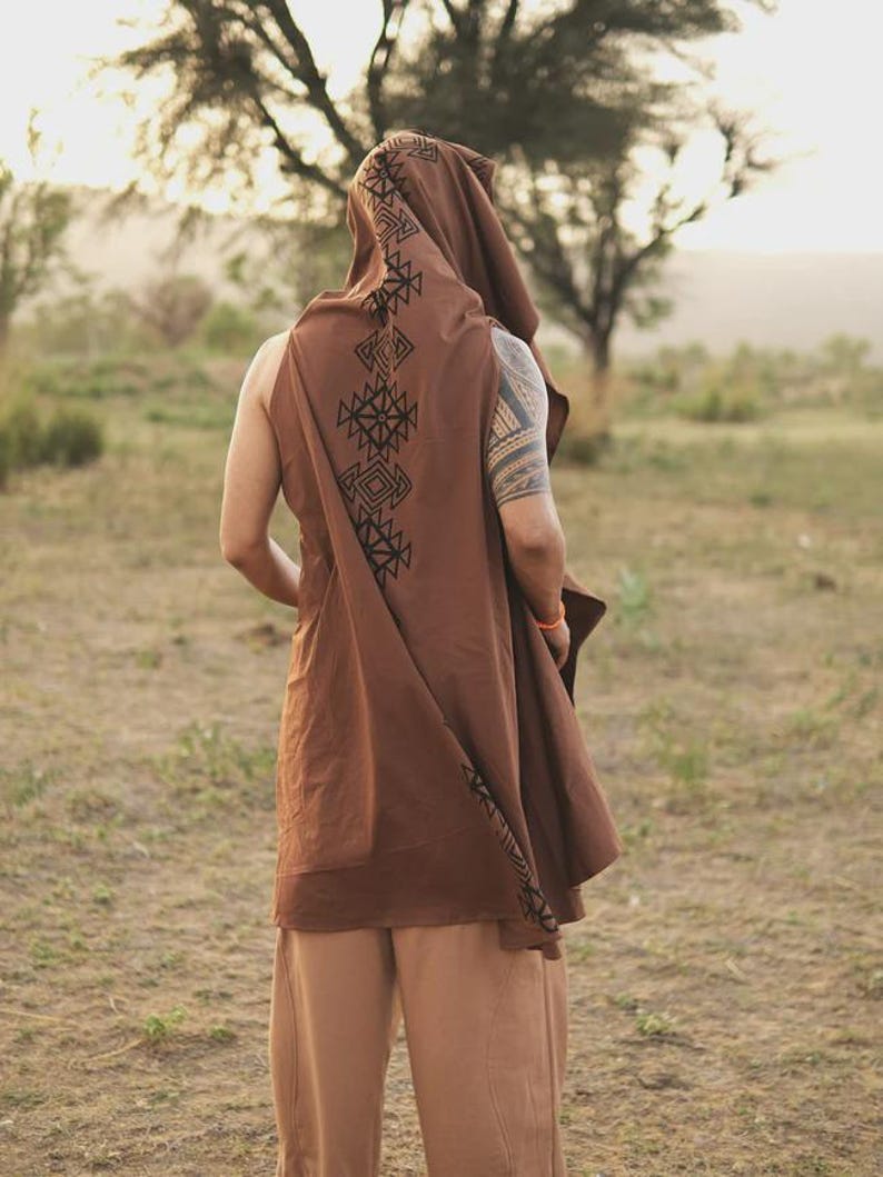 May include: A brown sleeveless hooded tunic featuring a black geometric pattern printed along the hood and down the back. The garment is worn with light brown trousers. The tunic has a loose, flowing fit and a pointed hood design.