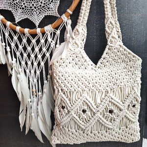 Puede incluir: Un bolso tote de macramé en color blanco roto con un intrincado patrón de nudos en forma de rombos y pequeñas cuentas de madera de color marrón oscuro integradas. El bolso tiene dos asas de hombro trenzadas y una estructura de cordón tejido.