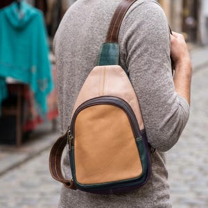 Könnte beinhalten: Ein mehrfarbiger Leder-Umhängerucksack mit einem hellbeigen oberen Bereich, einer beigen Vordertasche sowie dunkelgrünen und tiefvioletten Akzenten im unteren Teil. Der Rucksack verfügt über einen braunen Lederriemen und dunkle Metallreißverschlüsse.
