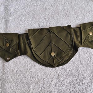 Può includere: Una cintura multiuso color verde oliva con tre tasche distinte caratterizzate da un motivo di cuciture a forma di foglia. Ogni tasca è fissata con un bottone a pressione in metallo color ottone. La cintura ha un design strutturato con bordi appuntiti.