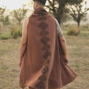 May include: A sleeveless brown garment worn over the shoulders, featuring a vertical row of black geometric patterns running down the center back. The fabric drapes loosely, extending past the waistline with a simple, open-front design.