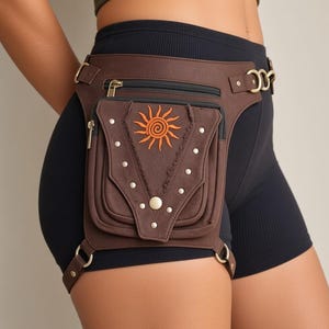 May include: A brown utility leg bag featuring a zipped main compartment and a front pocket adorned with an embroidered orange sun motif. The bag includes metallic silver studs, brass-toned hardware, and adjustable straps that secure around the waist and thigh.
