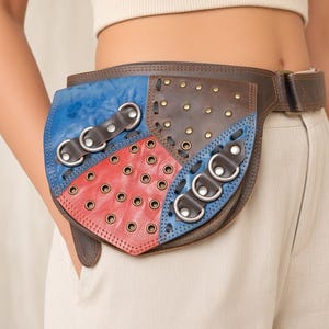 May include: A multi-colored leather waist bag featuring a patchwork design of blue, red, and dark brown panels. The bag is adorned with numerous metal grommets, decorative stitching, and silver-toned D-ring hardware attached to leather straps.