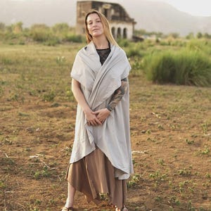 May include: A light gray sleeveless wrap-style vest with a draped collar and open front, layered over a black top and a brown midi-length skirt with an asymmetrical hemline, paired with simple brown sandals.