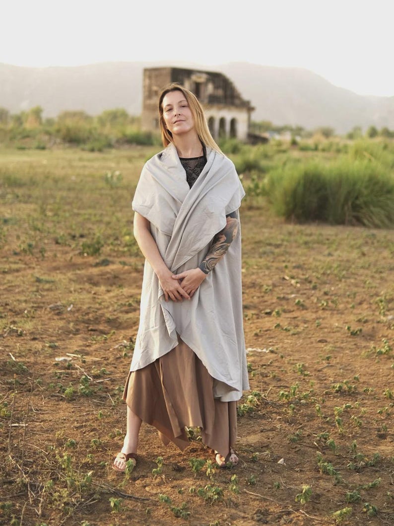May include: A light gray sleeveless wrap-style vest with a draped collar and open front, layered over a black top and a brown midi-length skirt with an asymmetrical hemline, paired with simple brown sandals.
