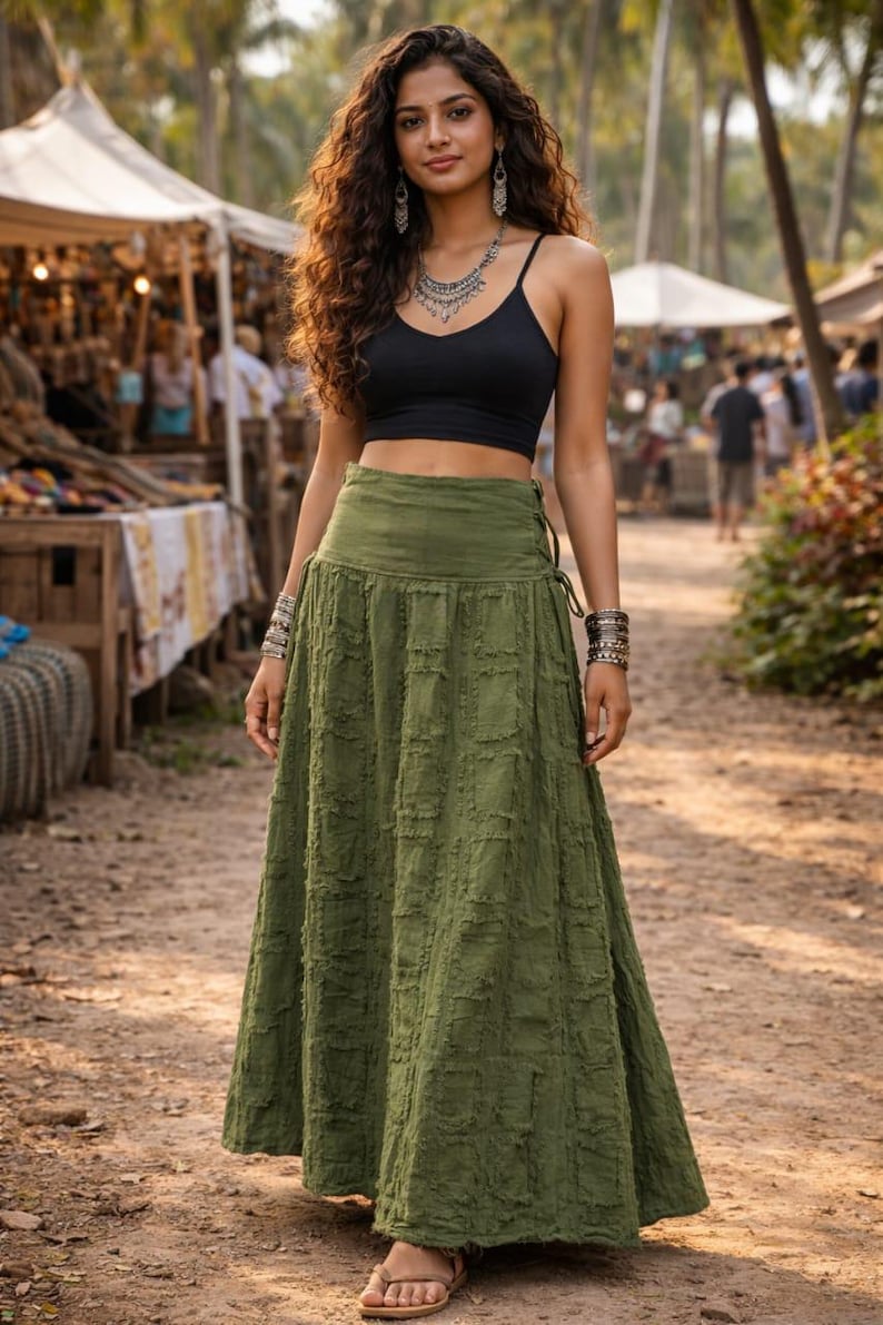 May include: A floor-length olive green skirt featuring a textured, grid-like pattern with frayed edges, a high waistband, and side lace-up detailing. The skirt is paired with a simple black spaghetti strap crop top and silver jewellery accessories.