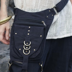 Puede incluir: Riñonera de lona negra con múltiples bolsillos y detalles metálicos dorados. La riñonera cuenta con un compartimento principal con cremallera, un bolsillo frontal con solapa con anillos decorativos y correas ajustables. El diseño incluye remaches y una trabilla.
