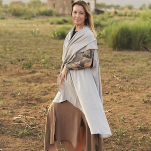 May include: A light gray wrap-style shawl worn over a black top and a brown skirt with a side slit. The shawl features a smooth, solid-colored fabric that drapes over the shoulders and down the torso, paired with brown thong-style sandals.