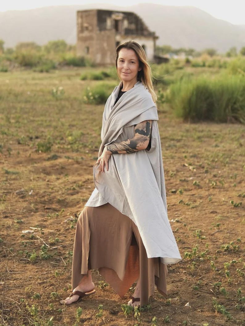 May include: A light gray wrap-style shawl worn over a black top and a brown skirt with a side slit. The shawl features a smooth, solid-colored fabric that drapes over the shoulders and down the torso, paired with brown thong-style sandals.