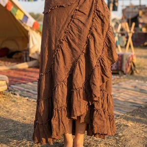 May include: A long, brown skirt with multiple ruffled layers. The skirt has a textured appearance and a wrap-style design. The model is standing barefoot on the ground, with a tent and colorful flags in the background.