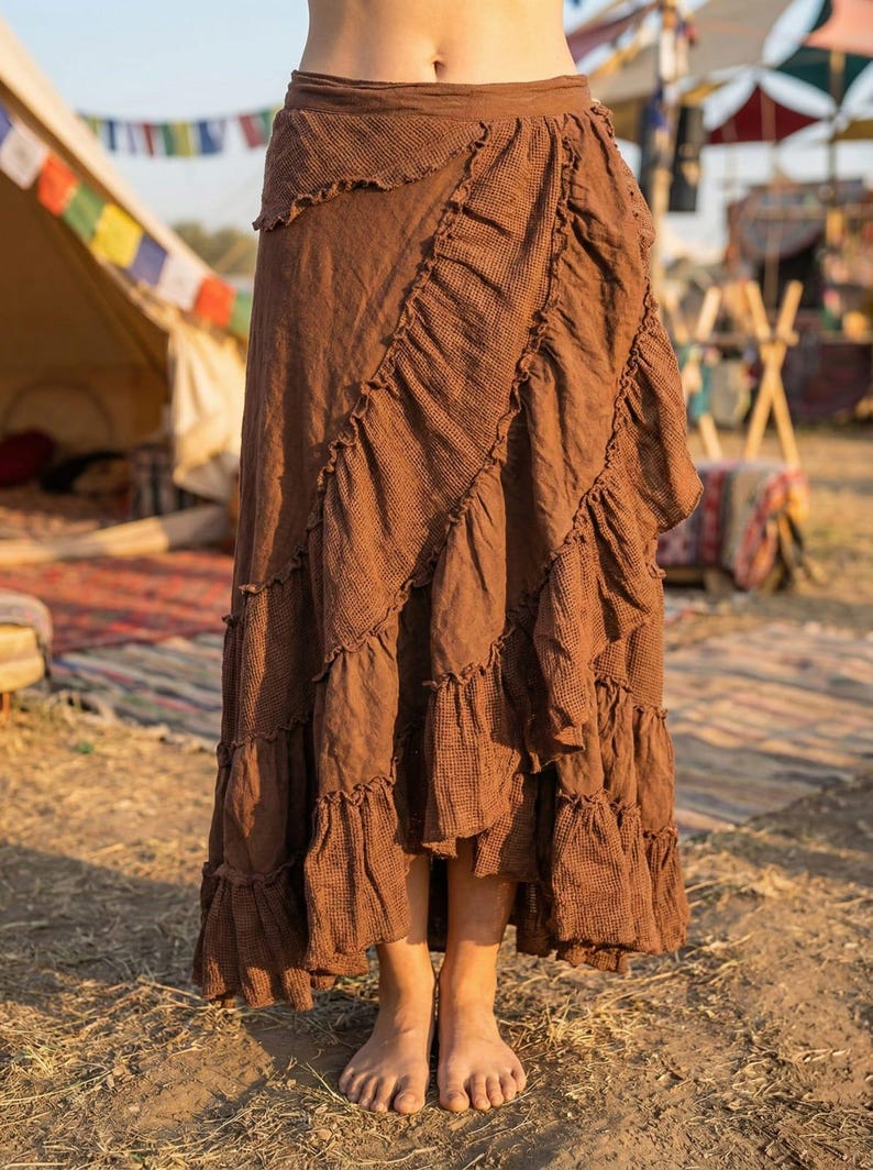 May include: A long, brown skirt with multiple ruffled layers. The skirt has a textured appearance and a wrap-style design. The model is standing barefoot on the ground, with a tent and colorful flags in the background.
