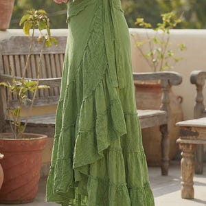 May include: A long, flowing green wrap skirt with a tiered, ruffled design. The skirt features a textured panel on the front and a tie closure at the waist. The skirt is made of a lightweight fabric and is suitable for warm weather.