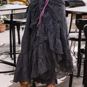 May include: A dark gray, layered skirt with lace detailing and a ruffled hem. The skirt is paired with brown leather sandals. A pink ribbon is tied at the waist. The skirt appears to be made of a soft, flowing fabric.