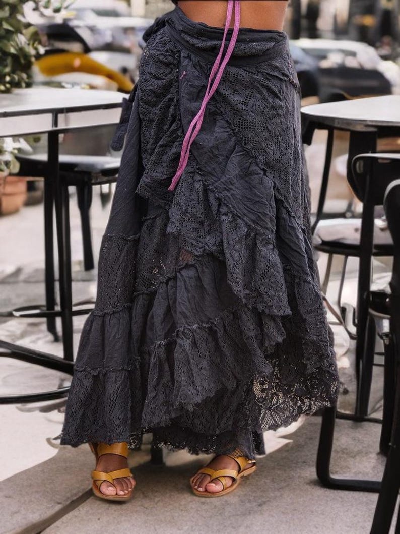 May include: A dark gray, layered skirt with lace detailing and a ruffled hem. The skirt is paired with brown leather sandals. A pink ribbon is tied at the waist. The skirt appears to be made of a soft, flowing fabric.
