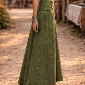May include: An olive green maxi skirt featuring a high-waisted design with a lace-up corset-style closure on the side. The fabric has a textured, repeating rectangular pattern throughout the length of the skirt, which flows down to the ankles.