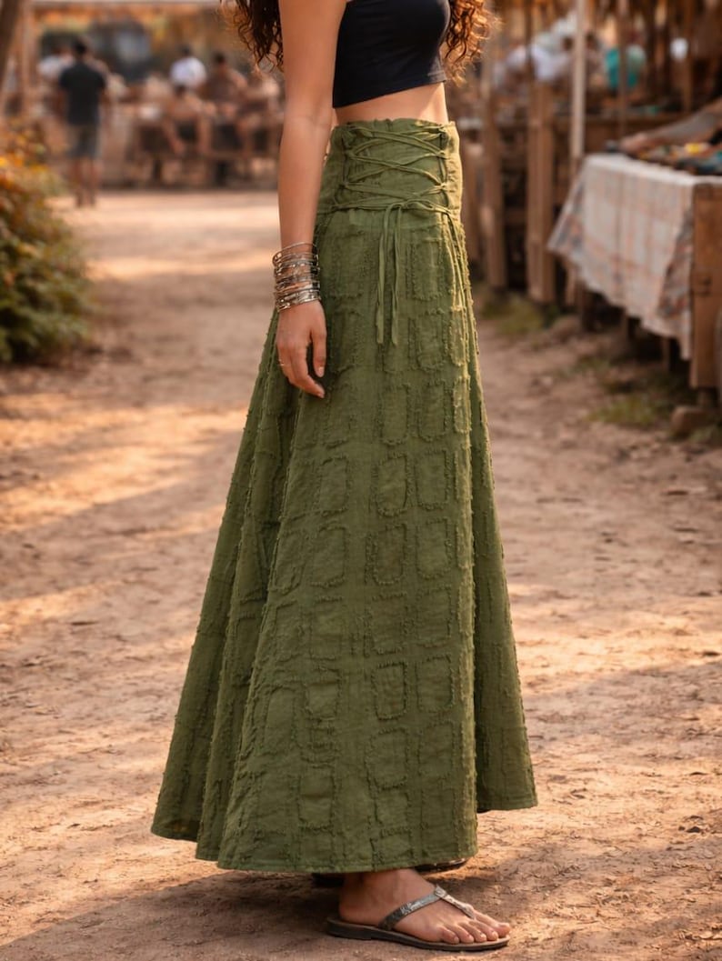 May include: An olive green maxi skirt featuring a high-waisted design with a lace-up corset-style closure on the side. The fabric has a textured, repeating rectangular pattern throughout the length of the skirt, which flows down to the ankles.