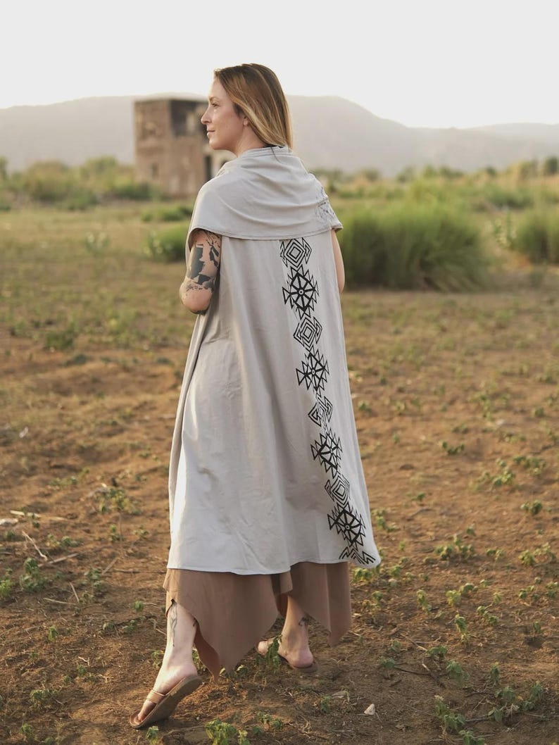 May include: A light gray sleeveless duster vest featuring a draped cowl neckline and a vertical column of black geometric diamond-shaped patterns printed down the center of the back. The garment has an open front and a long, flowing silhouette.