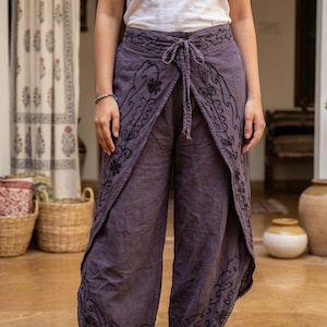 May include: Purple wrap-style pants featuring a white tank top. The pants have a tie-waist closure and are decorated with black embroidered floral patterns along the front panels and hemline, finished with a delicate black trim.