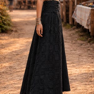 May include: A black maxi skirt featuring a high-waisted design with a lace-up corset-style closure at the waist. The fabric has a textured, repeating rectangular pattern throughout the length of the skirt, which falls to the ankles.