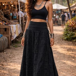 May include: A black sleeveless crop top with thin straps paired with a matching black high-waisted maxi skirt featuring a textured, distressed fabric finish. The outfit is accessorised with a silver-toned layered necklace, dangling earrings, and stacked metallic bracelets on both wrists.