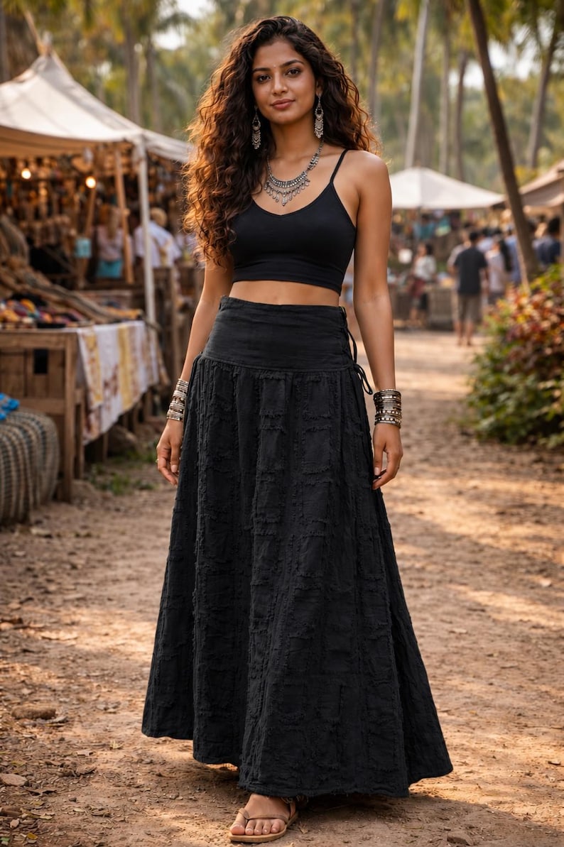 May include: A black sleeveless crop top with thin straps paired with a matching black high-waisted maxi skirt featuring a textured, distressed fabric finish. The outfit is accessorised with a silver-toned layered necklace, dangling earrings, and stacked metallic bracelets on both wrists.