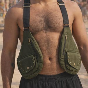 Puede incluir: Bolso de pecho verde oliva con dos bolsillos, cada uno con un bolsillo con cremallera y un bolsillo más pequeño con tachuelas de latón. El bolso tiene correas negras y herrajes metálicos dorados.