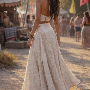 May include: A two-piece outfit consisting of a white crochet halter top and a matching long, textured off-white skirt. The skirt features a high-waisted design with side lace-up detailing and a frayed, layered fabric texture throughout the garment.