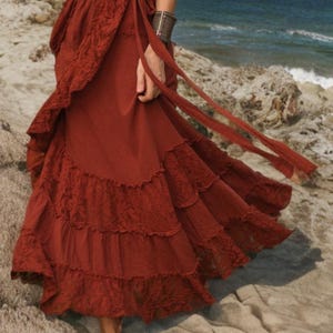 May include: A rust-colored wrap skirt with lace trim and tiered ruffles. The skirt has long ties and is worn near the ocean. The skirt is made of a flowing fabric.
