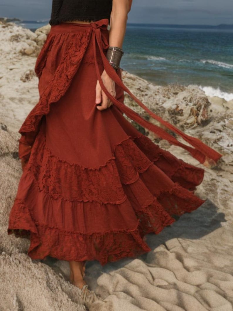 May include: A rust-colored wrap skirt with lace trim and tiered ruffles. The skirt has long ties and is worn near the ocean. The skirt is made of a flowing fabric.