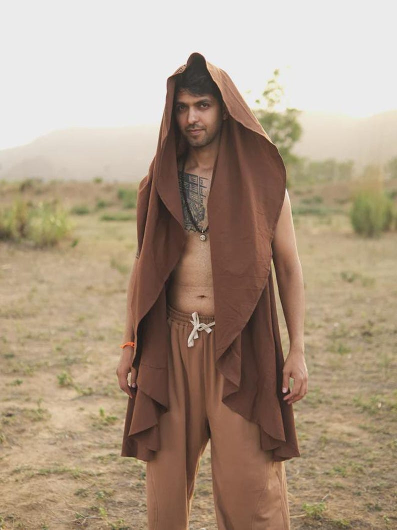 May include: A sleeveless brown hooded vest with an open front and draped fabric panels, paired with light brown drawstring sweatpants. The vest features a large, pointed hood and a flowing, layered design that extends down to the mid-thigh area.