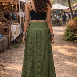 May include: A long olive green skirt featuring a textured square pattern with frayed edges and side lace-up details, paired with a black sleeveless crop top. The skirt reaches the ankles and has a wide waistband, worn with brown sandals.
