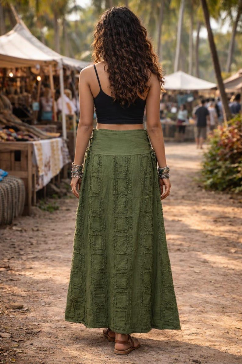 May include: A long olive green skirt featuring a textured square pattern with frayed edges and side lace-up details, paired with a black sleeveless crop top. The skirt reaches the ankles and has a wide waistband, worn with brown sandals.