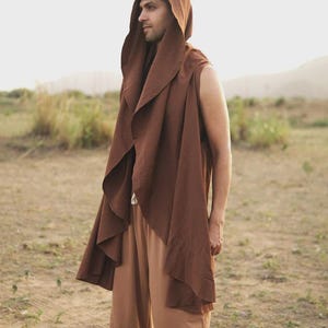 May include: A sleeveless brown hooded vest with an open front and draped fabric panels, paired with light brown trousers. The vest features a large, pointed hood and a long, flowing silhouette that extends past the waistline.