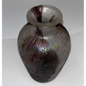 May include: A small, decorative glass vase with a rounded body and a wide, circular opening. The vase has a silver-gray iridescent finish with streaks of deep burgundy and hints of green and purple. The vase is likely used for displaying flowers or as a decorative item.
