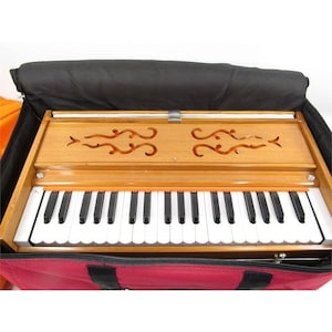 May include: A wooden harmonium musical instrument featuring a keyboard with black and white keys, a polished light brown wood finish, and decorative cut-out patterns on the top panel, all housed within a protective red and black carrying case.