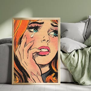 Puede incluir: Una impresión enmarcada de arte pop de una mujer llorando. La obra de arte presenta un primer plano del rostro de una mujer con cabello rojo brillante, ojos azules y lágrimas. La mujer se muerde el labio y su mano está cerca de su rostro.