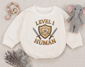 May include: A cream-colored long-sleeve baby bodysuit featuring a graphic of a shield with a twenty-sided die, two crossed swords, and the text "LEVEL 1 HUMAN" printed in brown. The garment is surrounded by a wooden bead teether and a gray elephant toy.