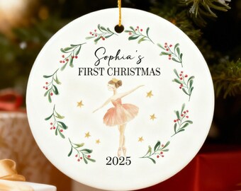 May include: A white ceramic Christmas ornament featuring a ballerina design. The ornament reads "Sophia's FIRST CHRISTMAS 2025" and is encircled by a wreath of green leaves and red berries. A gold ribbon is attached for hanging.